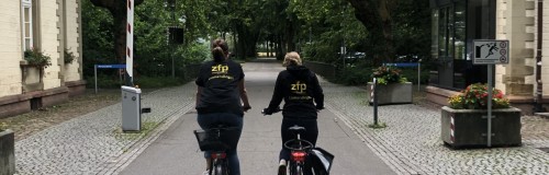 zwei Menschen fahren auf dem Fahrrad zwischen zwei Gebäuden durch Richtung Schranke