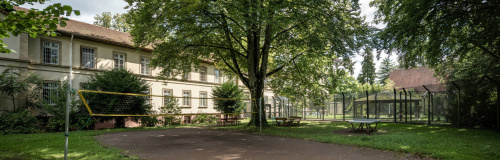 Gebäude der forensischen Klinik mit Bäumen und Volleyballfeld davor