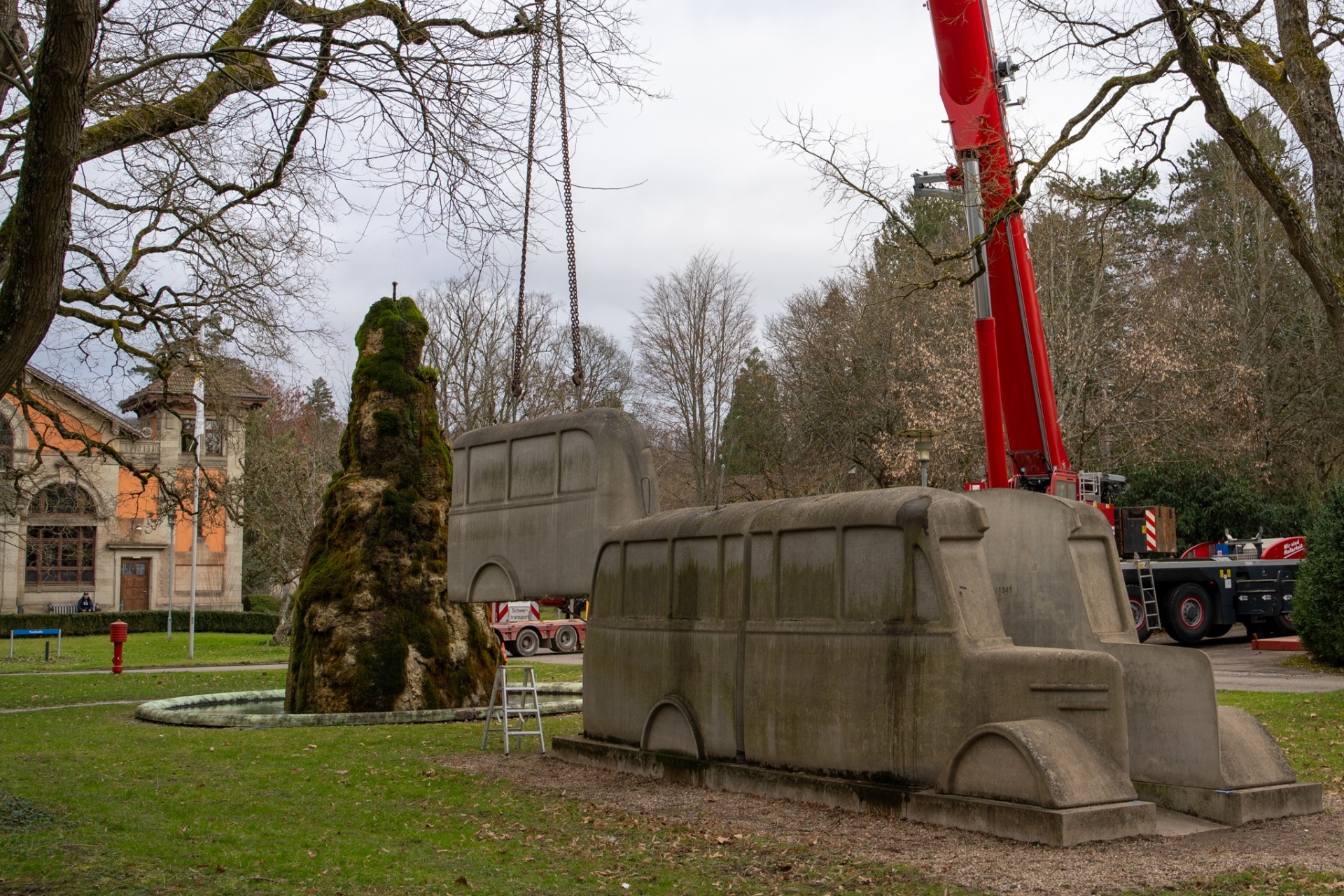 Denkmal Grauer Bus mit Kran und Brunnen im Hintergrund