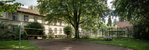 Gebäude der forensischen Klinik mit Bäumen und Volleyballfeld davor